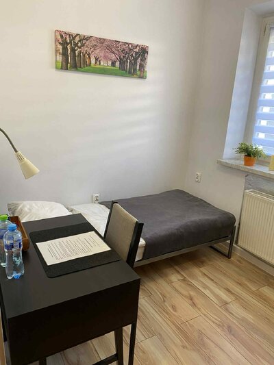 Wnętrze małego, jasnego pokoju z łóżkiem pojedynczym, biurkiem z lampką, oknem i obrazem kwitnących drzew na ścianie. Wnętrze małego, jasnego pokoju z łóżkiem pojedynczym, biurkiem z lampką, oknem i obrazem kwitnących drzew na ścianie.